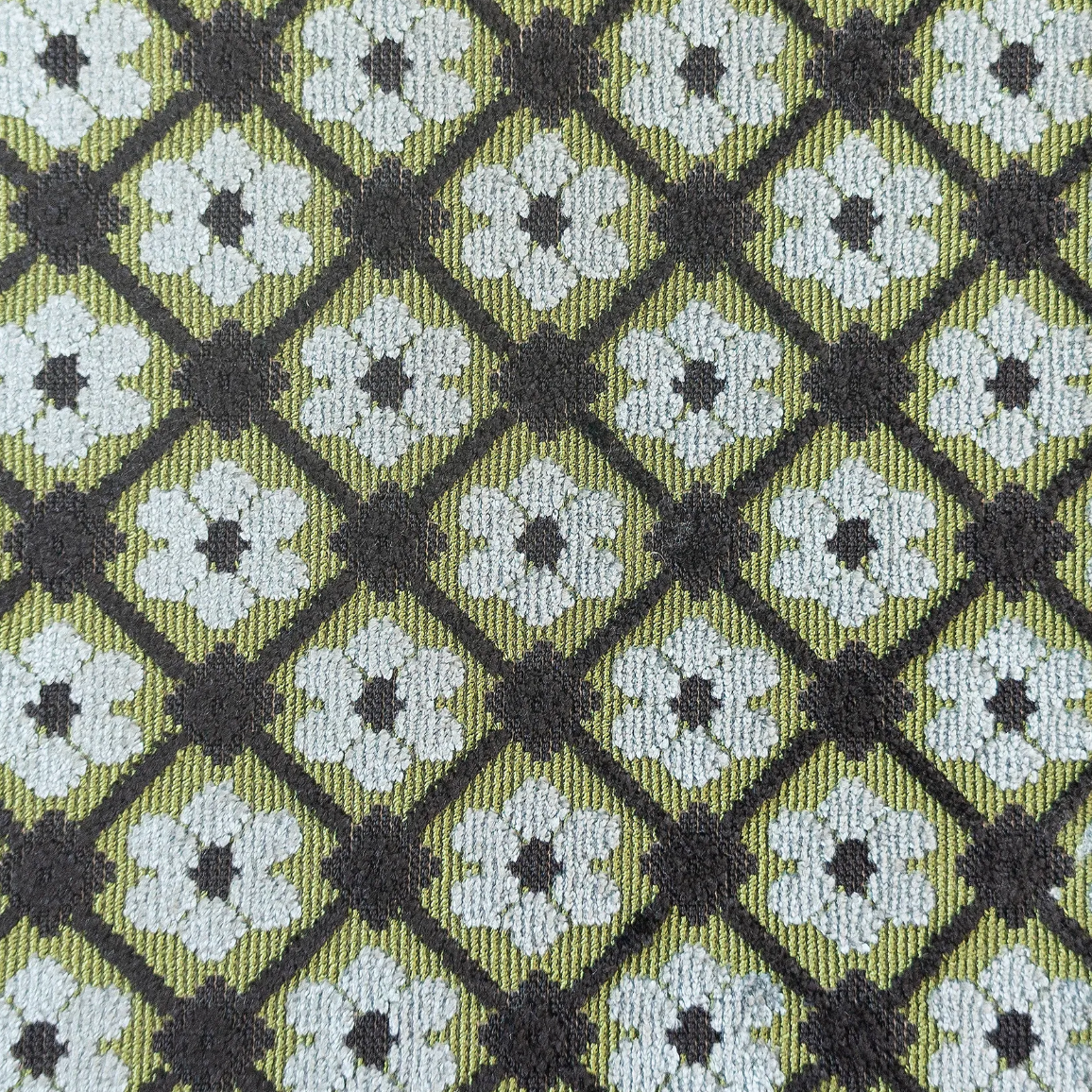 A close-up of a woven fabric with a repeating geometric pattern of white and dark gray floral motifs inside olive green diamonds.