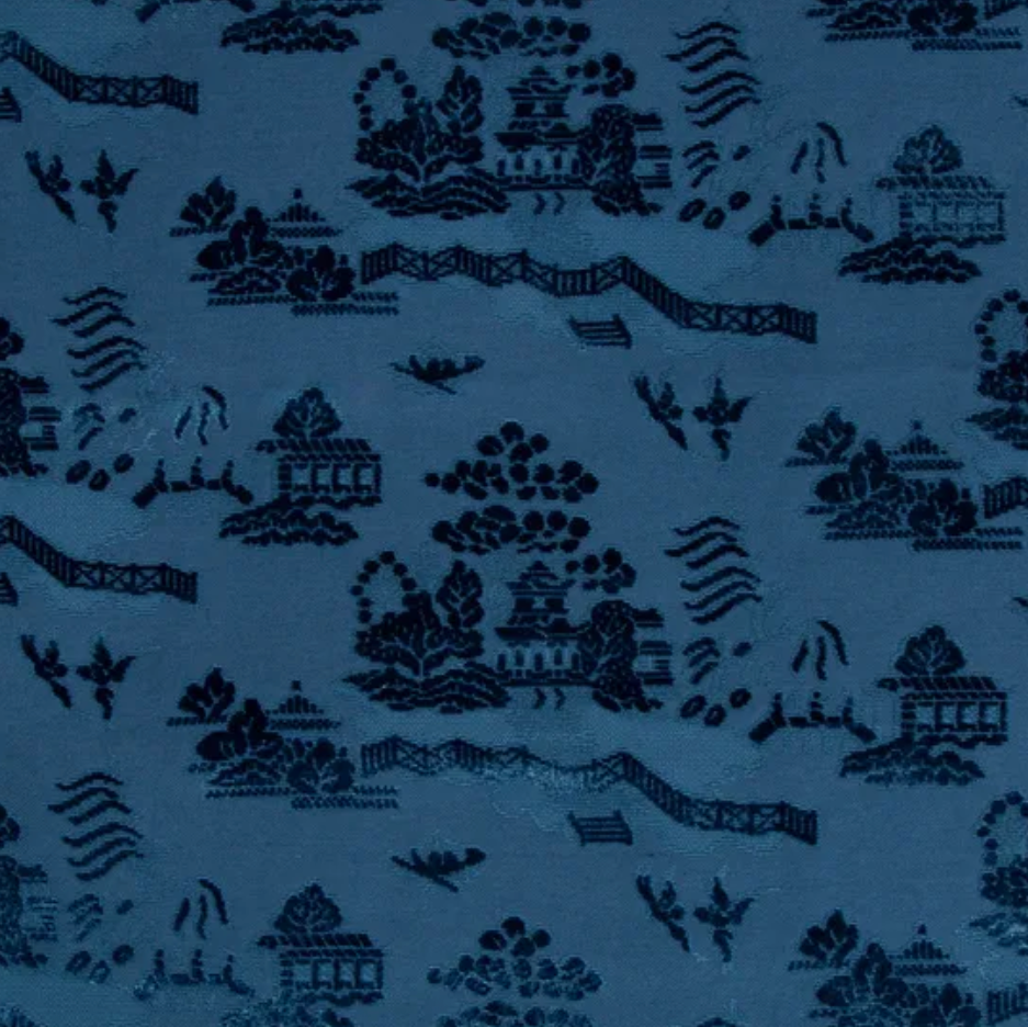 A close-up of a blue fabric with a dark navy chinoiserie pattern featuring pagodas, bridges, trees, and waves.